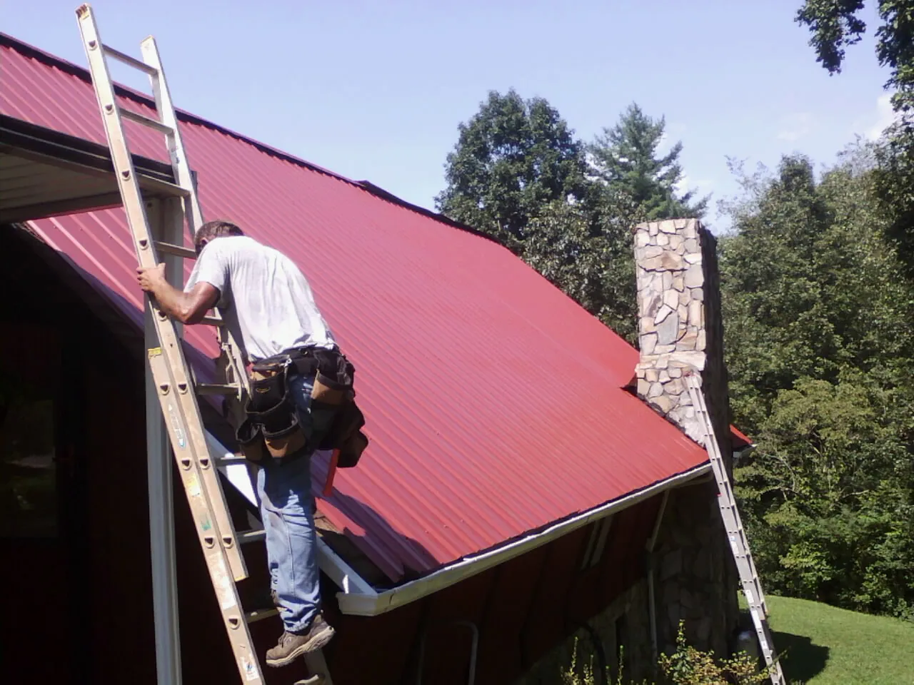 Roofer on a ladder beside a red metal roof and stone chimney — Soffit & Fascia Repair by Roofing Brothers in Fultonham